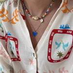 Woman wearing a colorful top and two necklaces. First is a small bead necklace with a tan fish bead at its center, second is a longer rectangle link gold chain with a blue enamel Heart pendant.