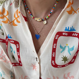 Woman wearing a colorful top and two necklaces. First is a small bead necklace with a tan fish bead at its center, second is a longer rectangle link gold chain with a blue enamel Heart pendant.