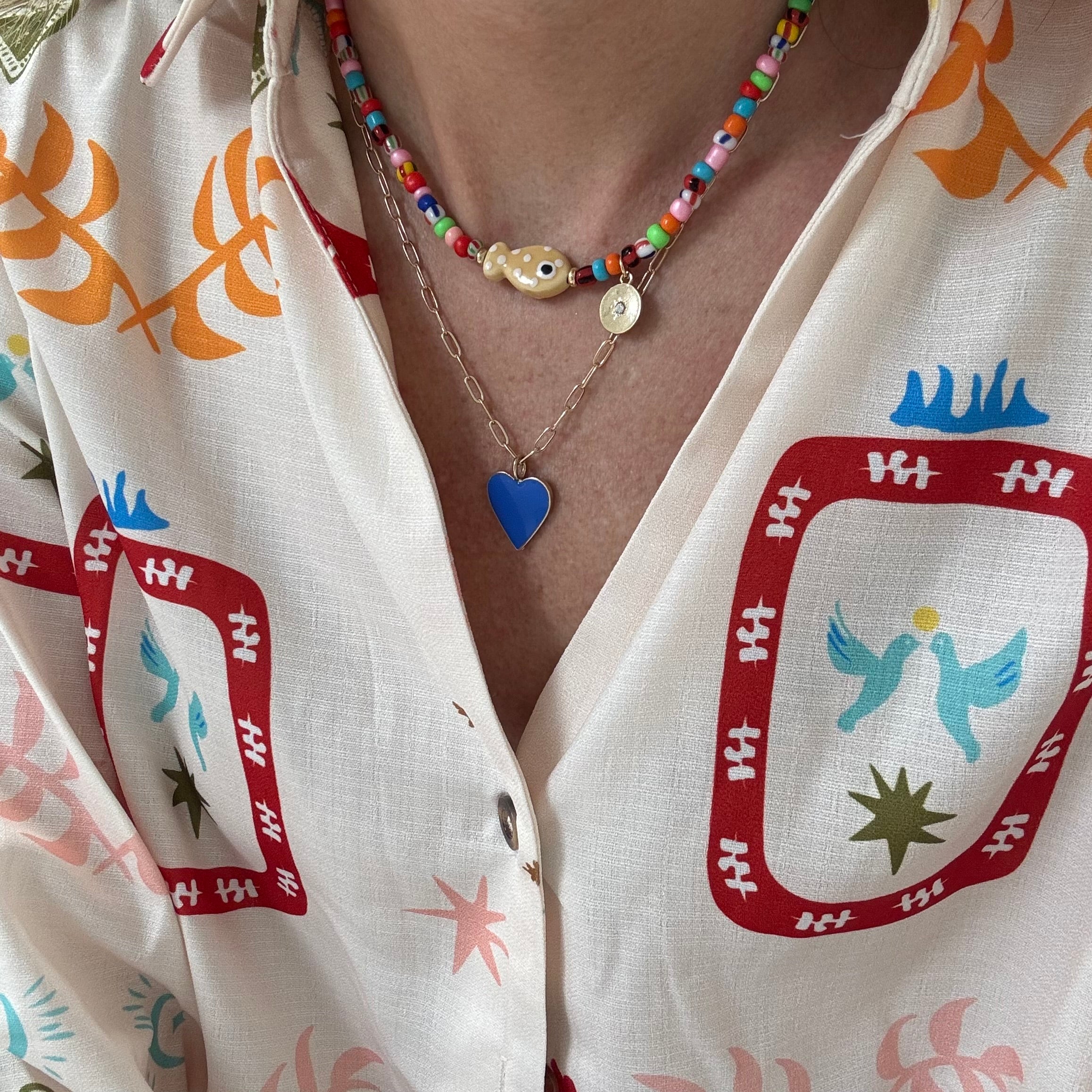 Woman wearing a colorful top and two necklaces. First is a small bead necklace with a tan fish bead at its center, second is a longer rectangle link gold chain with a blue enamel Heart pendant.