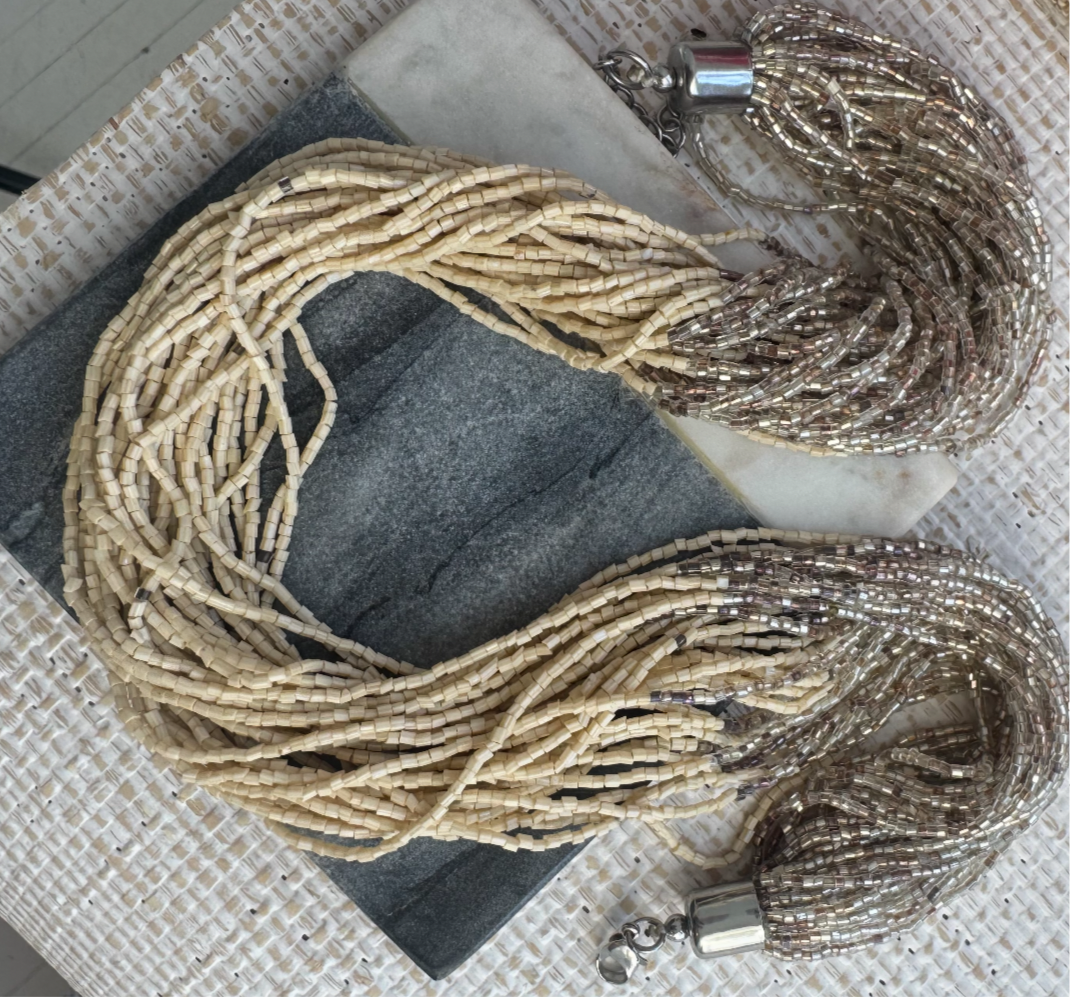 Picture of a statement necklace made of many strands of cream colored seed beads. Strands begin with clear tan beads, have a tan section in the middle and end with clear tan beads.