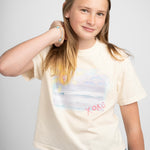 Picture of young girl wearing a cream colored Tshirt with a beach scene printed on the front with the letters XOXO in pink.