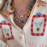 Woman wearing a brightly colored top. She is wearing three beaded necklaces. The first is a dainty white, gold and pink bead necklace, the second is a multicolored necklace with a tan fish and gold disc pendant and the last is a large bead necklace in blues, greens, yellows, pinks and oranges.