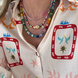 Woman wearing a brightly colored top. She is wearing three beaded necklaces. The first is a dainty white, gold and pink bead necklace, the second is a multicolored necklace with a tan fish and gold disc pendant and the last is a large bead necklace in blues, greens, yellows, pinks and oranges.