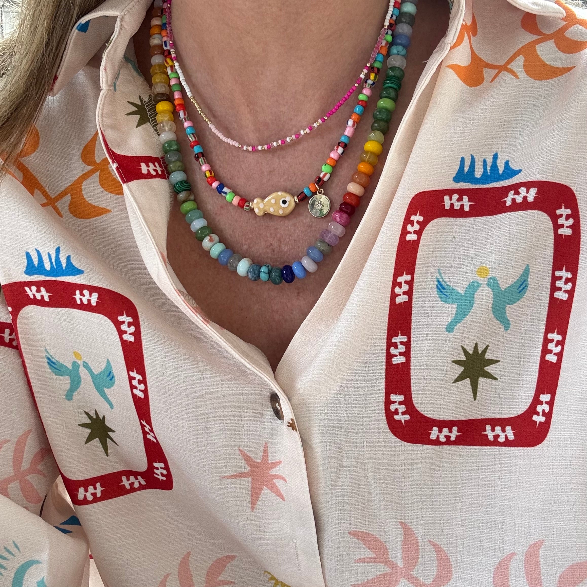 Woman wearing a brightly colored top. She is wearing three beaded necklaces. The first is a dainty white, gold and pink bead necklace, the second is a multicolored necklace with a tan fish and gold disc pendant and the last is a large bead necklace in blues, greens, yellows, pinks and oranges.