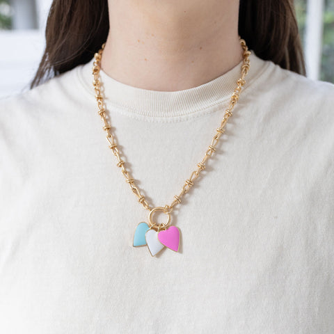 Model wearing gold toggle chain with blue, white and pink heart pendants.
