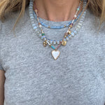 Picture of a woman wearing a gray bead necklace layered with a gold chain with a white enamel heart pendant and a smaller blue bead necklace.