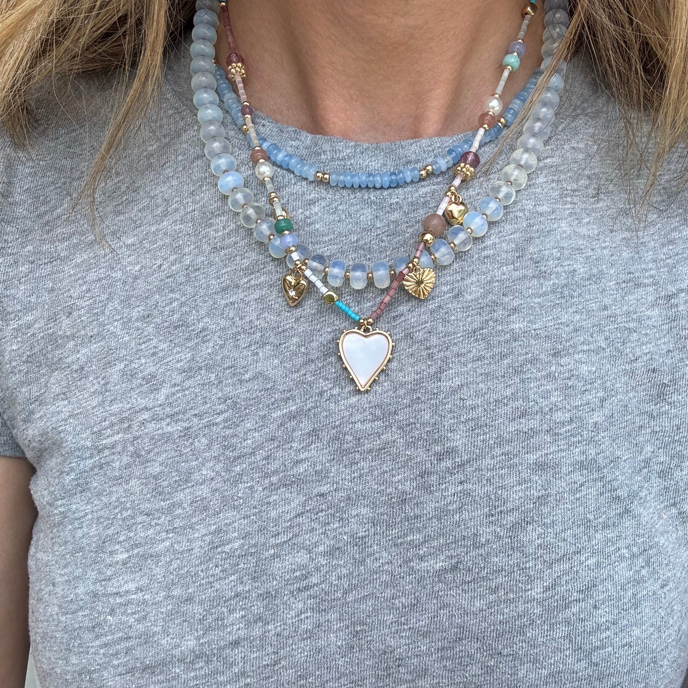 Picture of a woman wearing a gray bead necklace layered with a gold chain with a white enamel heart pendant and a smaller blue bead necklace.