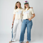 Picture of two women wearing jeans and matching beige cropped t shirts that have a beach scene.