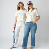 Picture of two women wearing jeans and matching beige cropped t shirts that have a beach scene.