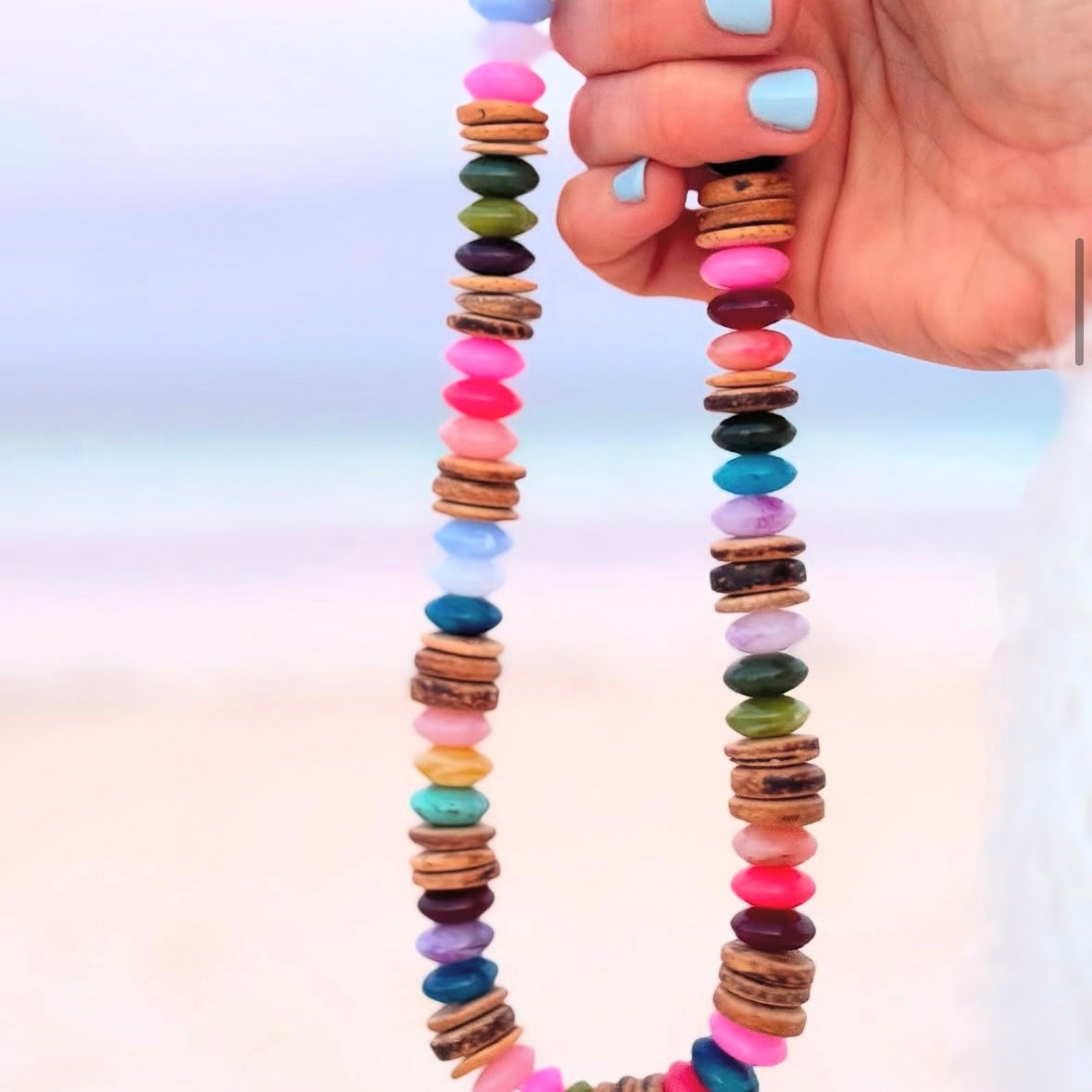 Picture of a hand holding a necklace made of multicolored beads and coconut shell beads. The necklace is made in mostly shades of pinks and blues.