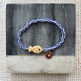 Picture of a bracelet made of blue and white striped beads. At its center is a tan fish bead with white polka dots and a small gold disc pendant.