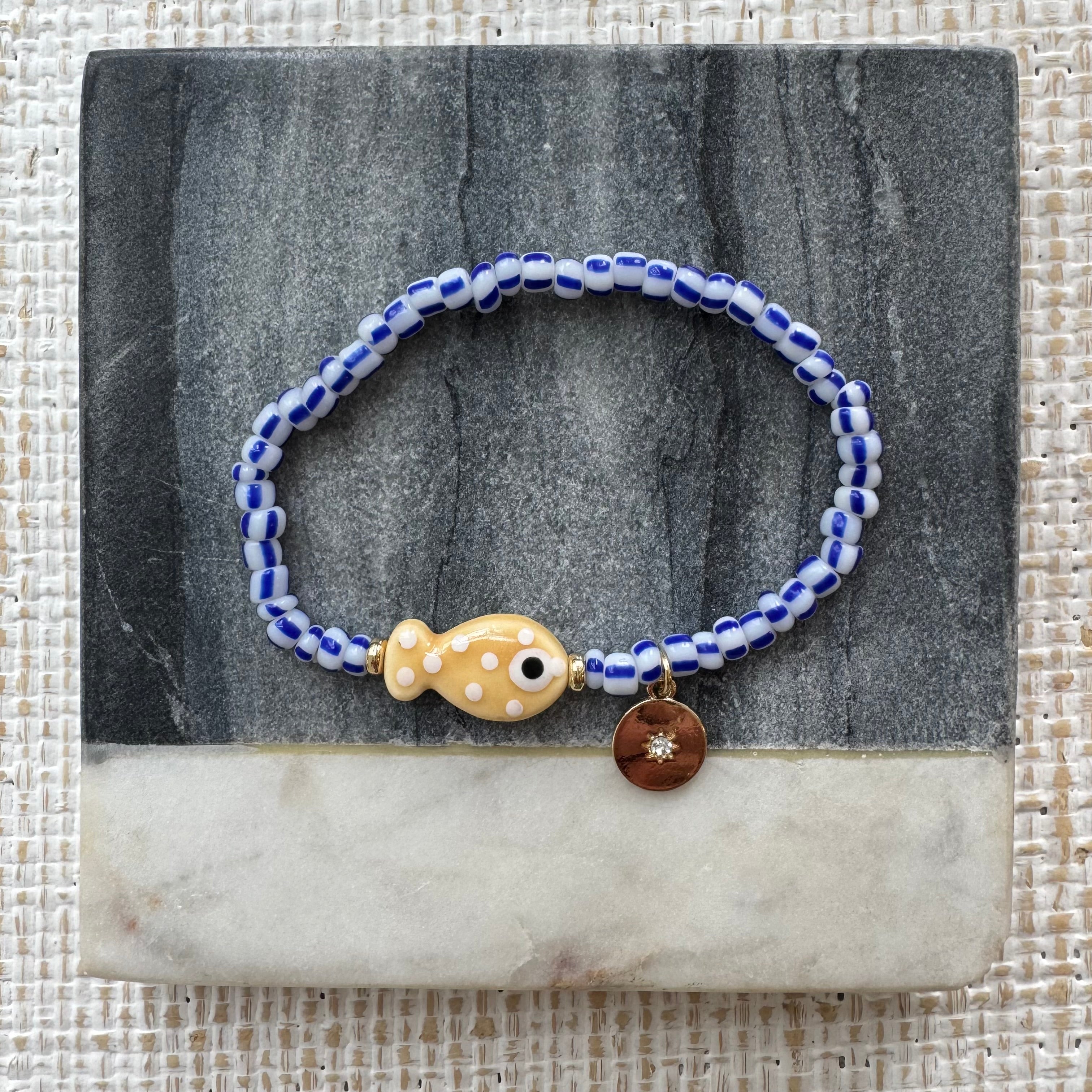 Picture of a bracelet made of blue and white striped beads. At its center is a tan fish bead with white polka dots and a small gold disc pendant.