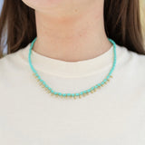 Model wearing a short necklace made of small turquoise beads and gold tear drop accent beads.
