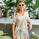Woman indoors by an island bar with plants behind her in a colorful dress wearing sunglasses and posing/smiling