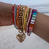 Picture of a woman's wrist wearing 17 different bracelets. 15 bracelets are tiny beaded bracelets of different color with gold accent beads. Bracelets are red, pink, blue, orange, tan, yellow and white. One thick linked gold wrap around bracelet with a large gold heart charm and one multicolored rectangular beaded bracelet.