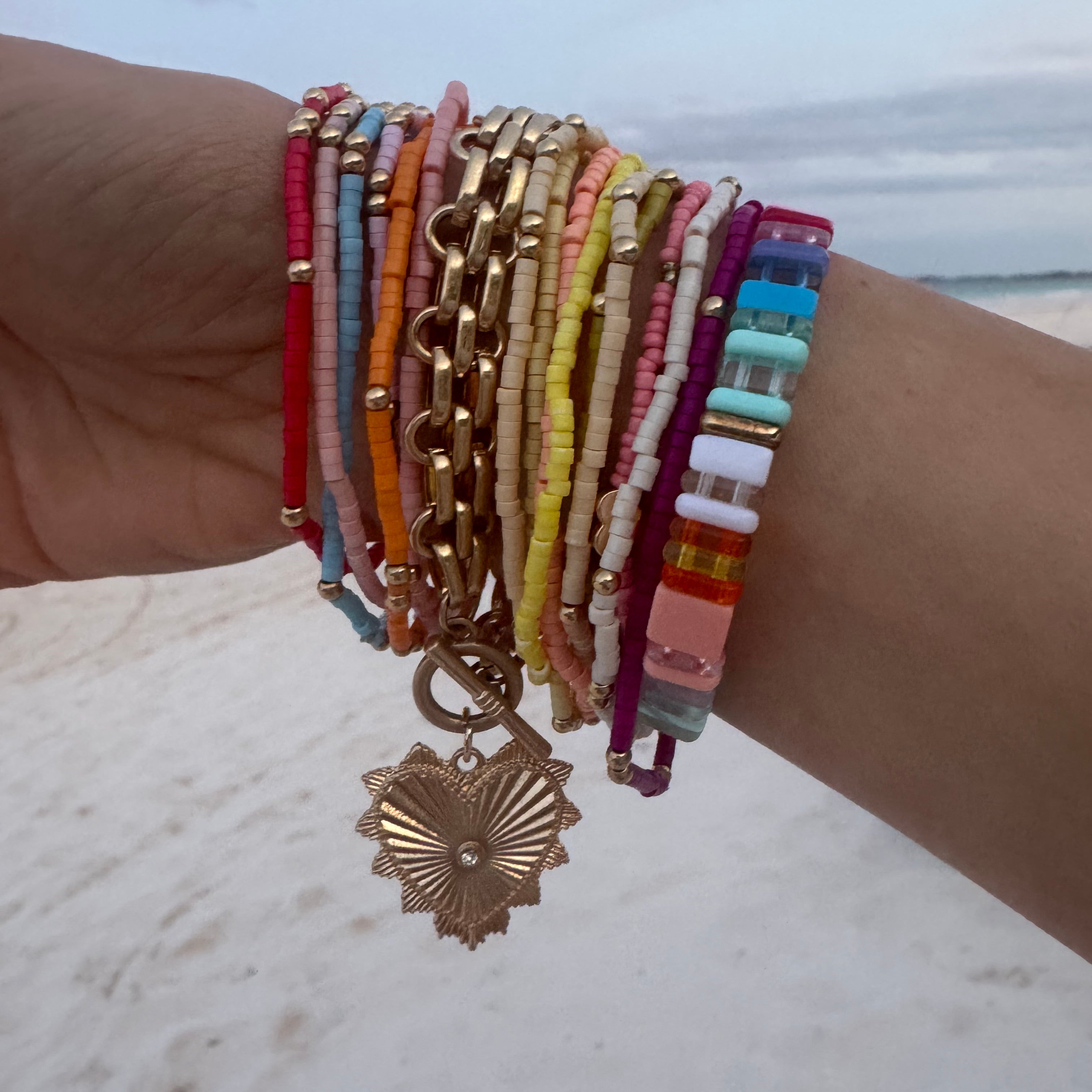 Picture of a woman's wrist wearing 17 different bracelets. 15 bracelets are tiny beaded bracelets of different color with gold accent beads. Bracelets are red, pink, blue, orange, tan, yellow and white. One thick linked gold wrap around bracelet with a large gold heart charm and one multicolored rectangular beaded bracelet.