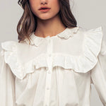 Picture of a model wearing a white long sleeved dress shirt. Shirt has a ruffle collar around the neck and breast and is a button up shirt.