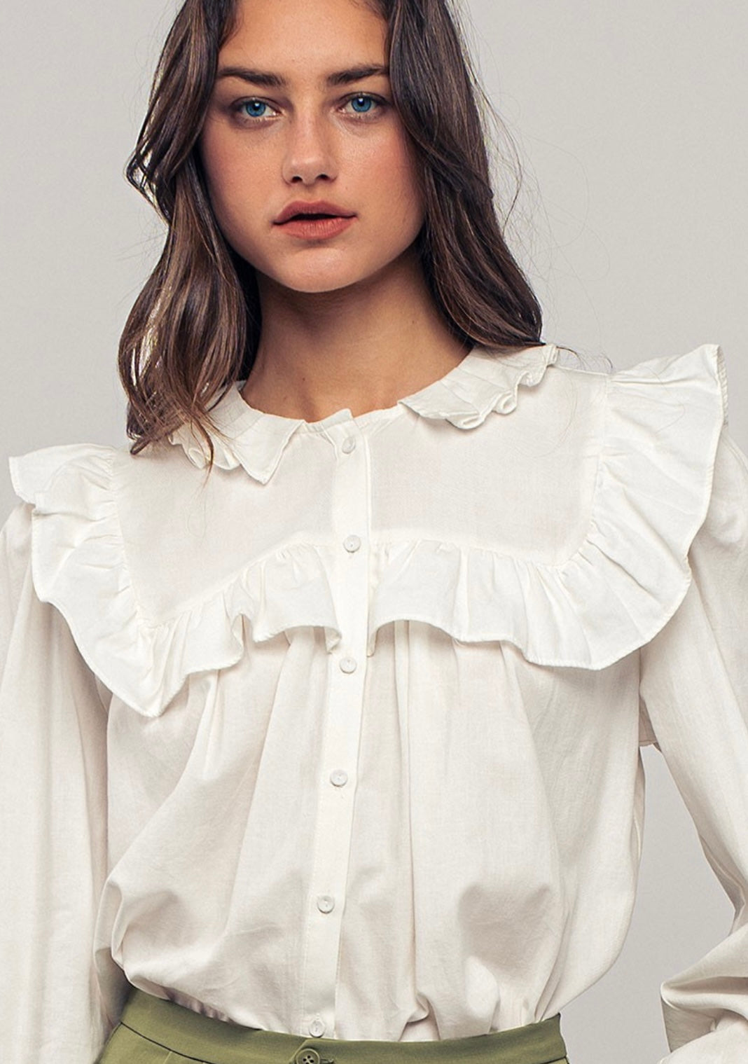 Picture of a model wearing a white long sleeved dress shirt. Shirt has a ruffle collar around the neck and breast and is a button up shirt.