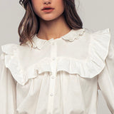 Picture of a model wearing a white long sleeved dress shirt. Shirt has a ruffle collar around the neck and breast and is a button up shirt.
