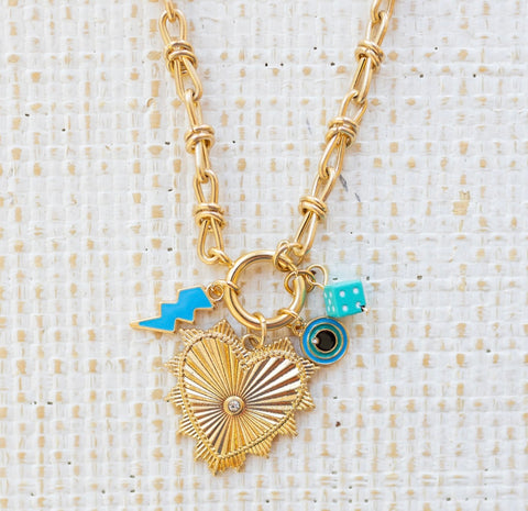 Picture of gold toggle necklace with a large heart pendant with a diamond at the center. Necklace has three charms attached. One a blue lightening bolt, an evil eye and a blue dice.