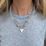 Picture of a woman wearing a gray bead necklace layered with a gold chain with a white enamel heart pendant and a smaller blue bead necklace.