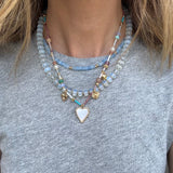 Picture of a woman wearing a gray bead necklace layered with a gold chain with a white enamel heart pendant and a smaller blue bead necklace.
