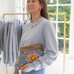 Woman standing in a house posing wearing a gray sweatshirt with blue stitching that says  "Joie De Vivre"