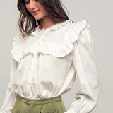 Picture of a model wearing a white long sleeved dress shirt. Shirt has a ruffle collar around the neck and breast and is a button up shirt.