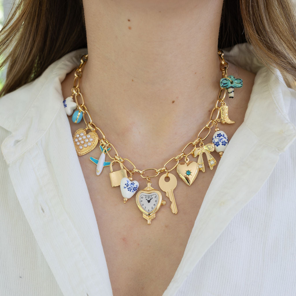 Picture of a gold charm chain necklace with 13 different charms.  Charms include a white star, gold heart, airplane, lock, heart, heart clock, key, gold heart with a blue diamond center, gold bow, ceramic white bead, Cowboy boot and palm tree.