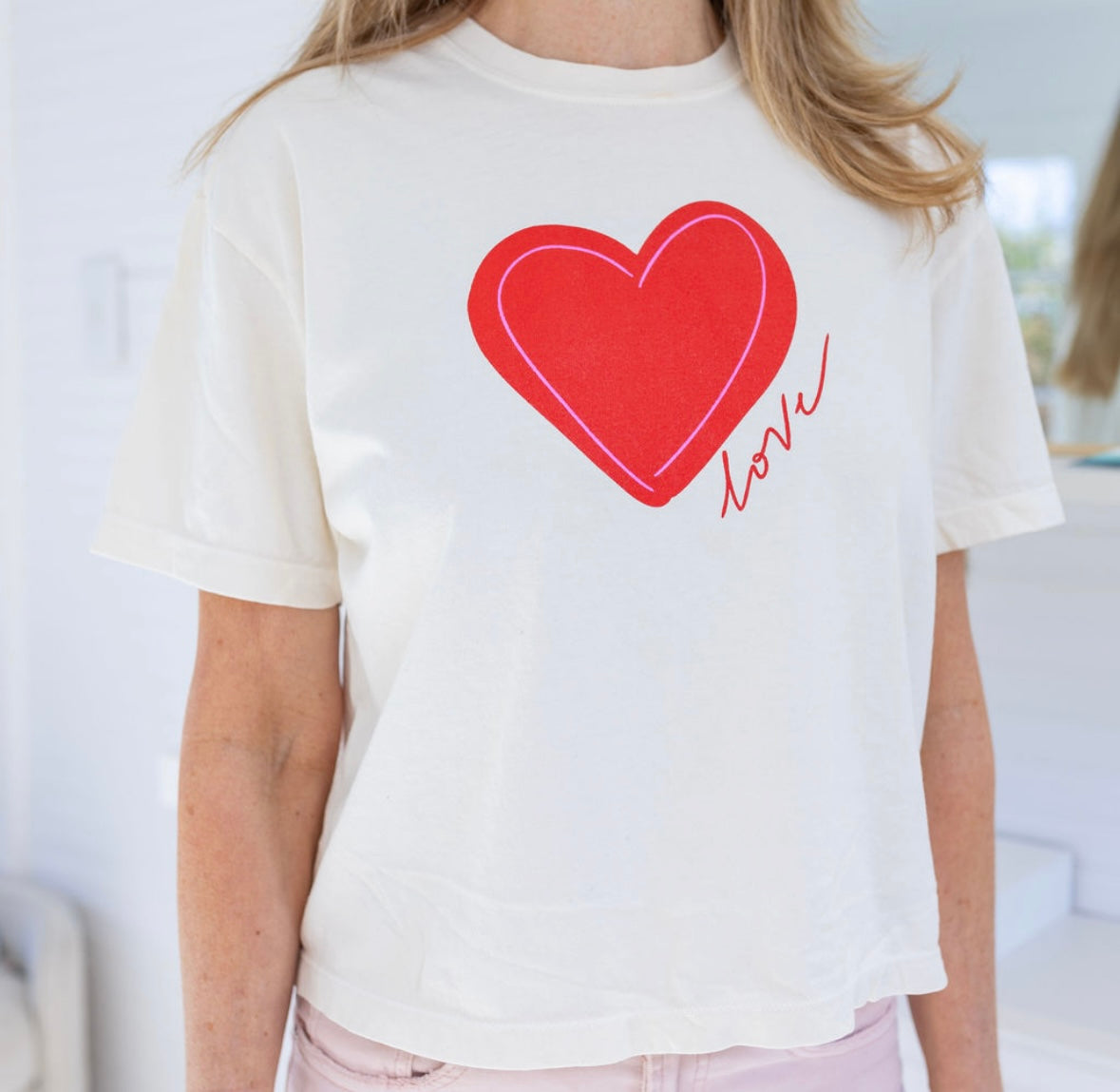 Woman wearing a a beige Tshirt with a red heart and script that reads "love".