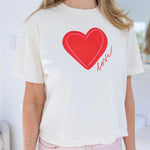 Woman wearing a a beige Tshirt with a red heart and script that reads "love".