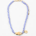 Picture of a bead necklace made of white and blue striped beads and a tan fish bead with a gold disc pendant.
