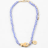Picture of a bead necklace made of white and blue striped beads and a tan fish bead with a gold disc pendant.