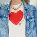 Woman wearing a denim jacket with embroidered hearts and a cream colored undershirt with a large red heart and red script that reads "love". Model is also wearing a colorful bead necklace and gold chain necklace with a heart pendant.