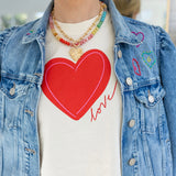 Woman wearing a denim jacket with embroidered hearts and a cream colored undershirt with a large red heart and red script that reads "love". Model is also wearing a colorful bead necklace and gold chain necklace with a heart pendant.