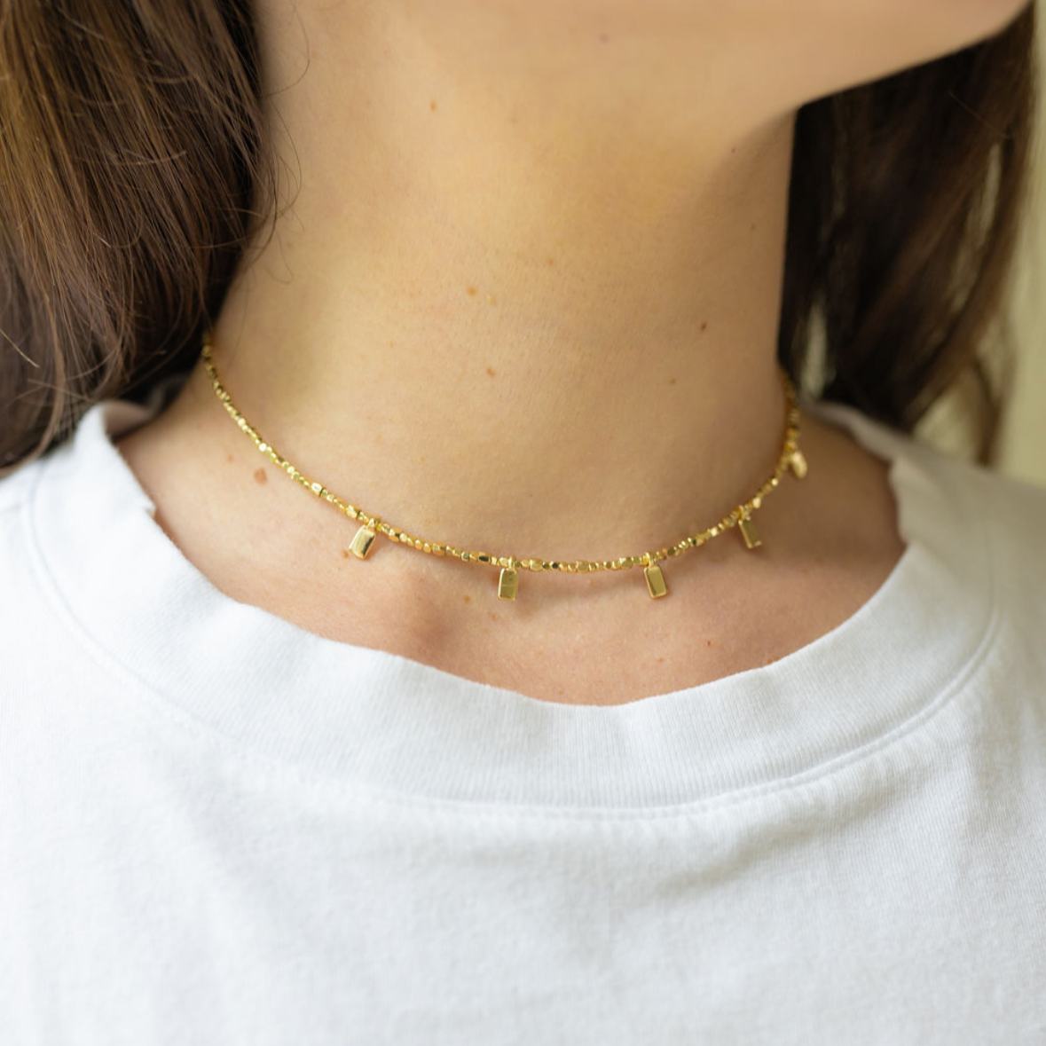 Girl wearing a gold necklace with a thin chain and little gold rectangles attached to it around her neck