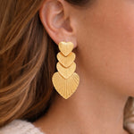 Gold, textured heart-shaped earrings worn by a person with brown hair.