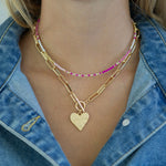 Gold chain link necklace with a gold heart pendant and toggle clasp and a dainty pink and white beaded necklace worn by a person with a denim jacket