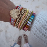 Picture of a woman's wrist wearing 17 different bracelets. 15 bracelets are tiny beaded bracelets of different color with gold accent beads. Bracelets are red, pink, blue, orange, tan, yellow and white. One thick linked gold wrap around bracelet with a large gold heart charm and one multicolored rectangular beaded bracelet.