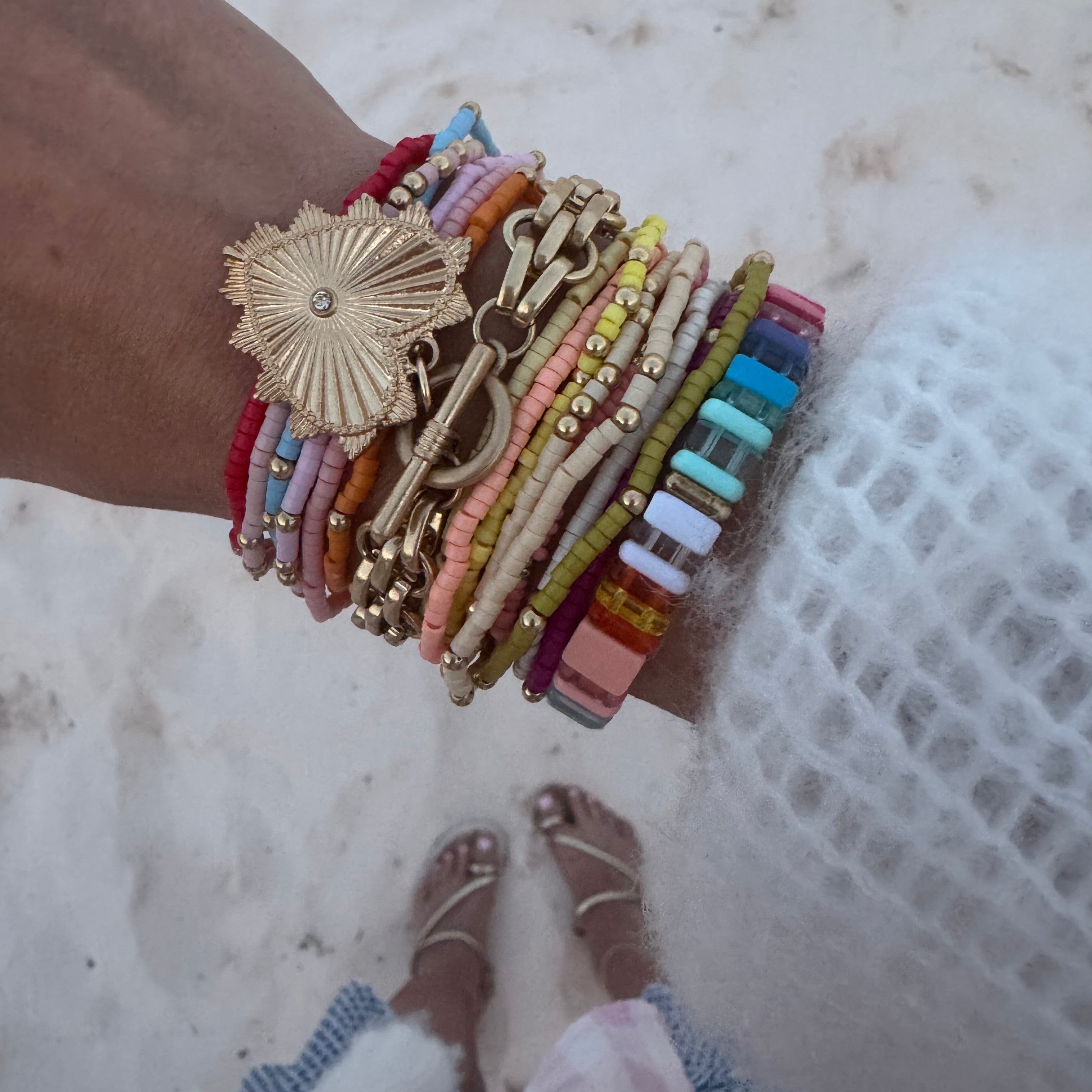 Picture of a woman's wrist wearing 17 different bracelets. 15 bracelets are tiny beaded bracelets of different color with gold accent beads. Bracelets are red, pink, blue, orange, tan, yellow and white. One thick linked gold wrap around bracelet with a large gold heart charm and one multicolored rectangular beaded bracelet.