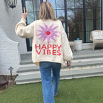 Picture of a woman walking away, holding up her hand in a peace sign, wearing an oversized cream colored sweater with a large purple sunburst with a red center and the words Happy Vibes in red at the bottom.