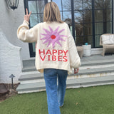 Picture of a woman walking away, holding up her hand in a peace sign, wearing an oversized cream colored sweater with a large purple sunburst with a red center and the words Happy Vibes in red at the bottom.
