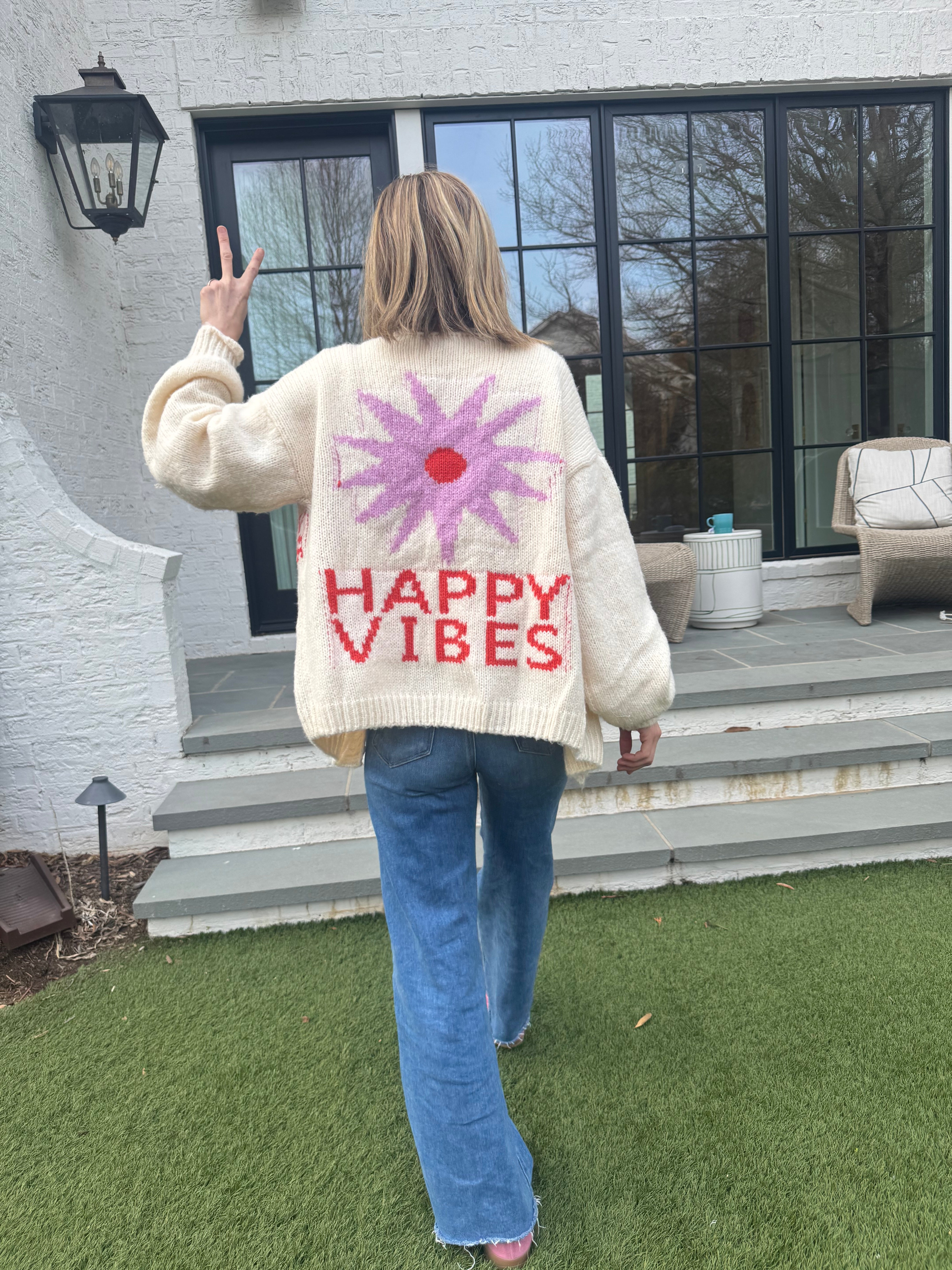 Picture of a woman walking away, holding up her hand in a peace sign, wearing an oversized cream colored sweater with a large purple sunburst with a red center and the words Happy Vibes in red at the bottom.