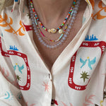 Woman wearing a colorful top and three necklaces. First necklace is a brightly colored bead necklace with a fish and small disc pendant. Second is a dainty multicolored bead necklace and the last is a larger clear bead necklace.