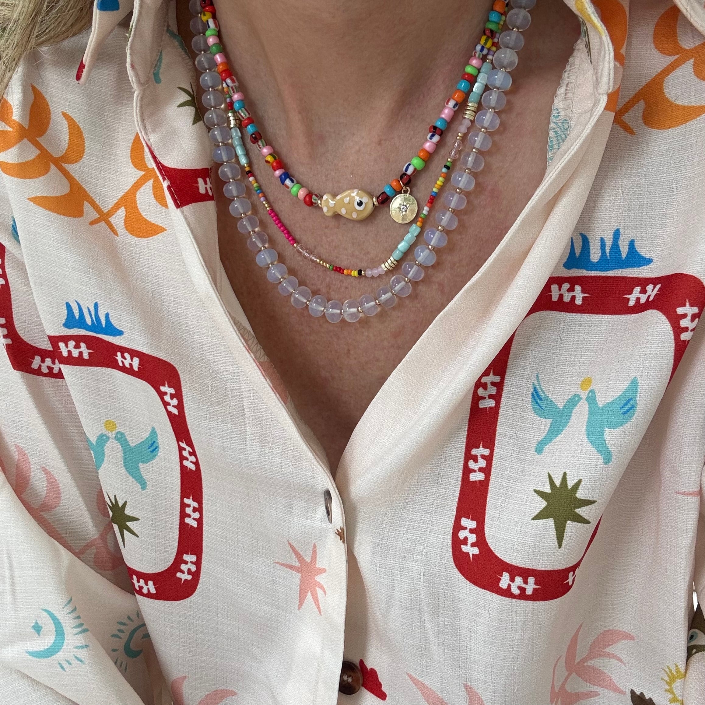 Woman wearing a colorful top and three necklaces. First necklace is a brightly colored bead necklace with a fish and small disc pendant. Second is a dainty multicolored bead necklace and the last is a larger clear bead necklace.