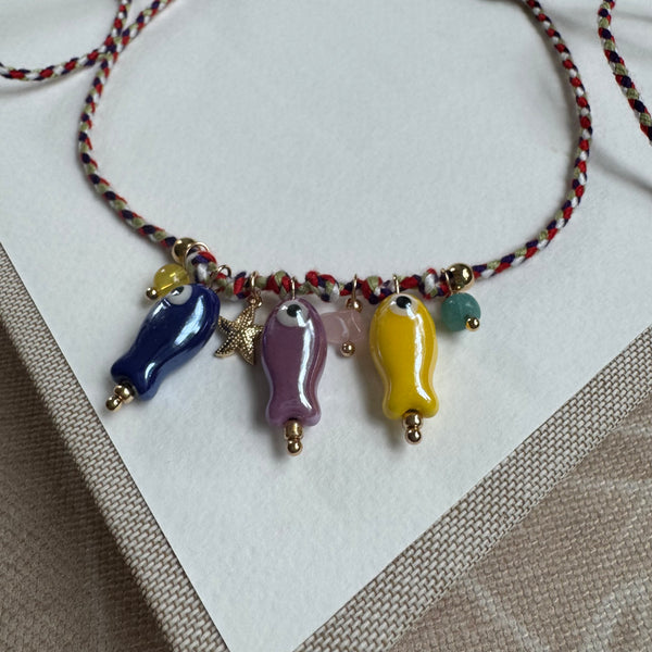 Picture of a thin woven string bracelet in red, white and black. Dangling from it are three fish beads in blue, purple and yellow with small yellow, pink and green accent beads and a small gold starfish.