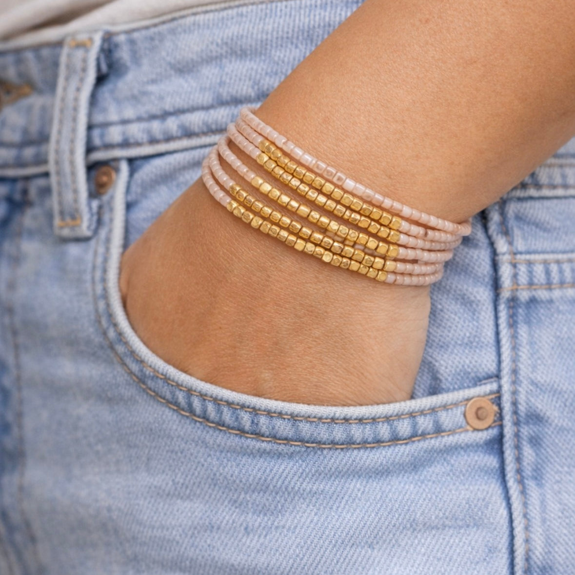 Close-up of a person wearing multiple light pink and gold beaded bracelets on a denim background