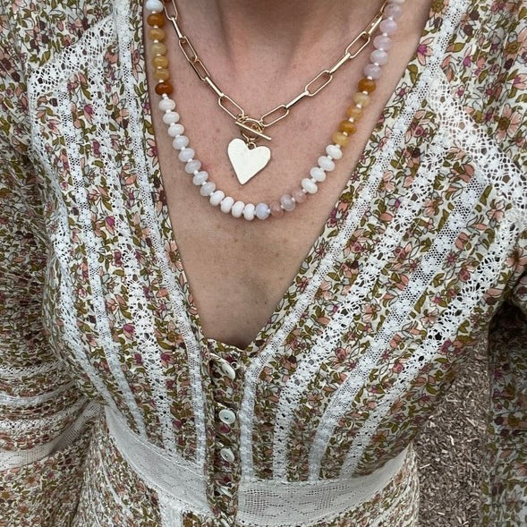 Picture of a woman's chest and dress wearing a set of stacked necklaces. One necklace has white beads and amber beads and one necklace with large gold links, a toggle clasp and with a gold heart charm.
