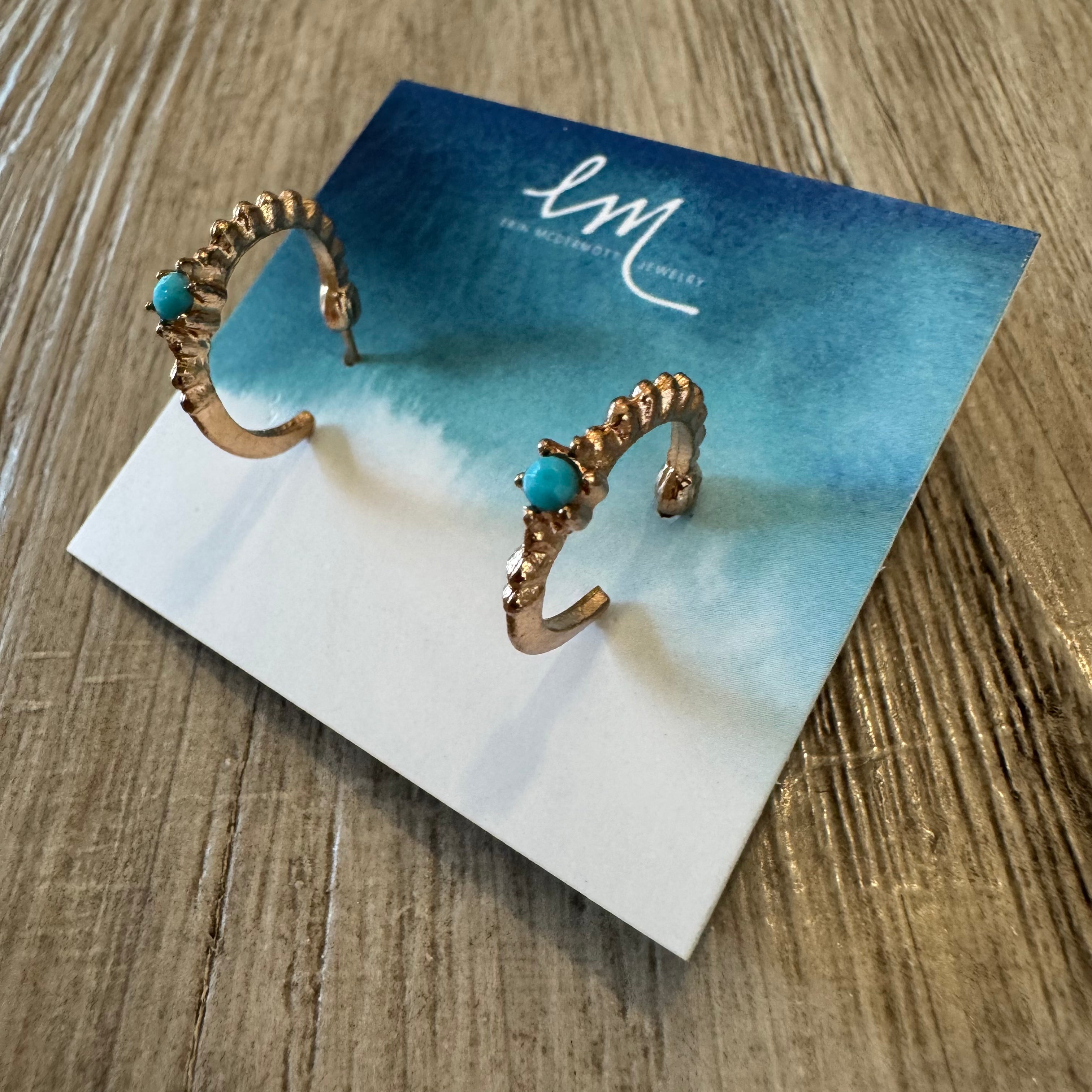 Gold earring hoops with turquoise stones on a branded card, placed on a wooden surface.