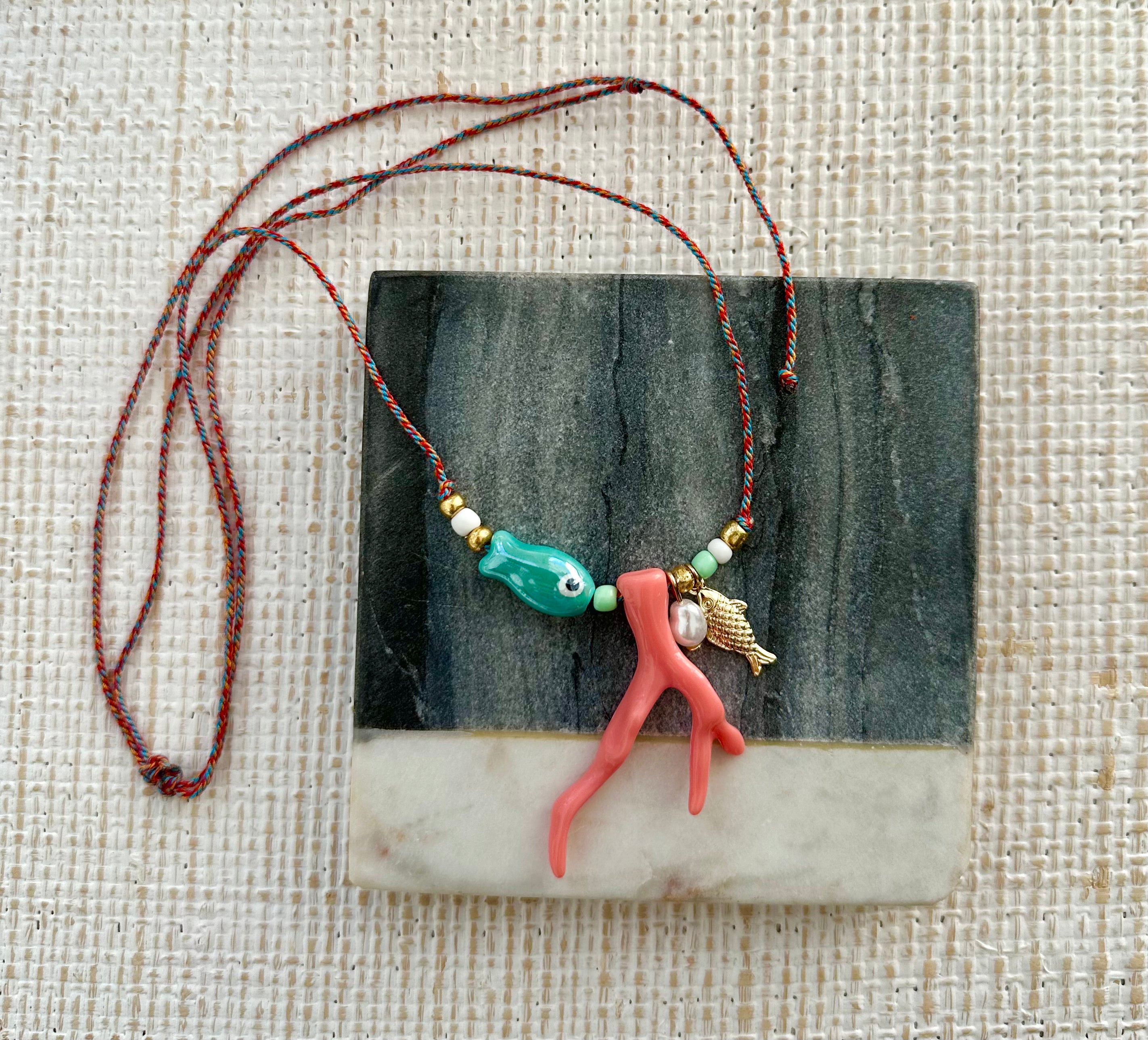 Picture of a woven string necklace with ocean themed beads on it. Main beads are a blue fish bead, a coral coral, a small freshwater Pearl pendant and a small gold fish pendant.